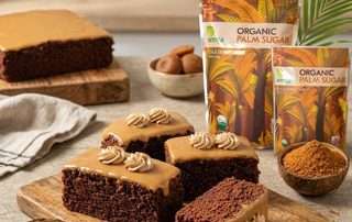 Foto beberapa potong kue cokelat tebal dengan lapisan karamel keemasan dan hiasan krim di atas talenan kayu. Di sampingnya terdapat produk kemasan "Arenga Organic Palm Sugar", batok kelapa berisi kelapa parut putih, dan batok kelapa berisi gula aren bubuk dengan latar belakang dapur yang hangat.