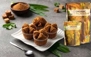 Empat buah kue mangkok gula aren klasik berwarna cokelat yang mekar sempurna, disajikan di atas piring keramik putih bergelombang. Di sampingnya terdapat produk kemasan Organic Palm Sugar Arenga, semangkuk gula aren bubuk, beberapa potongan gula aren cetak, sebatang sendok kecil, dan hiasan daun pandan segar di atas meja abu-abu bertekstur.