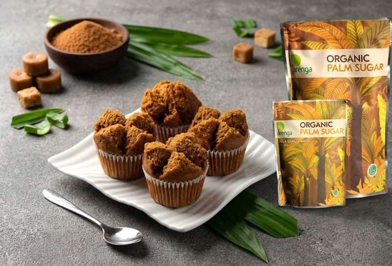 Empat buah kue mangkok gula aren klasik berwarna cokelat yang mekar sempurna, disajikan di atas piring keramik putih bergelombang. Di sampingnya terdapat produk kemasan Organic Palm Sugar Arenga, semangkuk gula aren bubuk, beberapa potongan gula aren cetak, sebatang sendok kecil, dan hiasan daun pandan segar di atas meja abu-abu bertekstur.