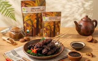 Foto premium food photography Sate Sapi Manis yang mengilat di atas piring tanah liat beralaskan daun pisang. Di latar belakang, terdapat kemasan kembar Arenga Organic Palm Sugar, stoples kaca berisi gula semut, teko tanah liat tradisional, dan mangkuk saus kecil di atas meja kayu dengan pencahayaan hangat.