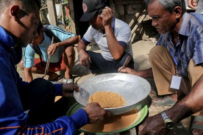 Empat orang pria perajin sedang berkumpul mengamati dengan saksama proses penyaringan bubuk gula semut menggunakan saringan logam lebar. Kolaborasi dan edukasi pengolahan yang efisien ini merupakan bagian dari upaya menekan dampak lingkungan dari produksi gula aren.