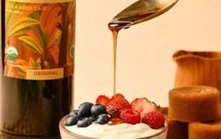 Sajian Resep Dessert Sehat: Greek Yogurt Beri Segar Penggugah Selera di dalam mangkuk keramik dengan taburan buah blueberry dan stroberi, disiram perlahan menggunakan gula aren cair dari atas sendok, berlatar belakang botol Arenga Organic Liquid Palm Sugar dan kepingan gula aren di atas alas anyaman.