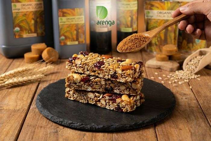 Tumpukan tiga potong granola bar bertekstur padat yang terbuat dari campuran oat, kacang almond, kacang mete, biji bunga matahari, dan cranberry kering di atas alas hitam. Di bagian atas terdapat sendok kayu berisi bubuk pemanis berwarna cokelat, merepresentasikan aplikasi gula aren dalam kuliner moderen.