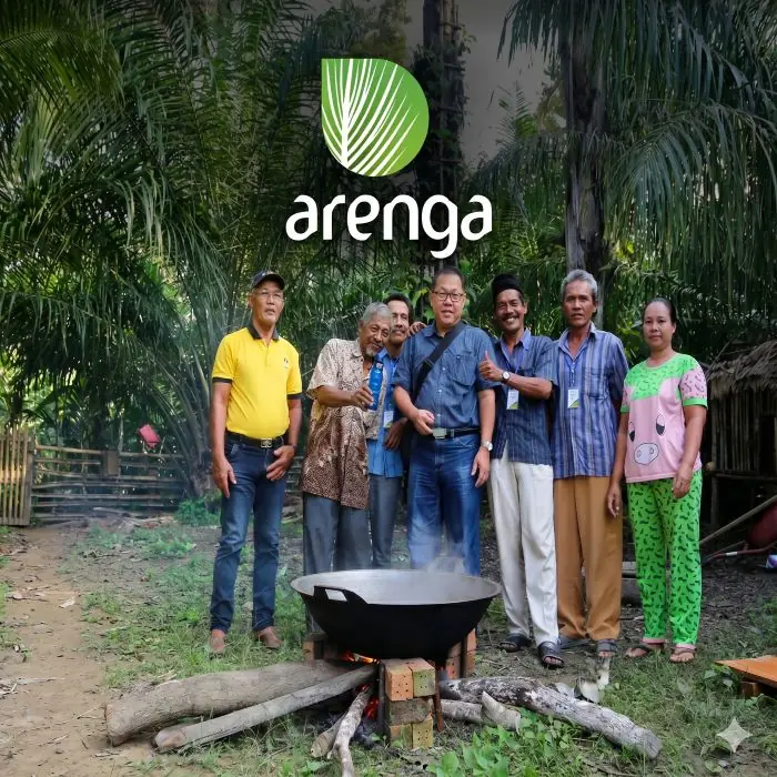 produksi gula aren berkelanjutan Sebuah foto kelompok delapan orang (tujuh pria dan satu wanita) dengan berbagai pakaian, berdiri bersama di tengah hutan pohon aren yang lebat di jalan setapak tanah. Mereka berkumpul mengelilingi kuali besar mengepul di atas tungku api kayu sederhana. Latar belakang menunjukkan pepohonan aren yang rimbun dan gubuk kayu. Di bagian tengah atas gambar terdapat logo melingkar daun aren hijau Arenga dengan teks 'arenga' berwarna putih. Seorang pria memegang botol nira biru dan pria lainnya memberikan acungan jempol.
