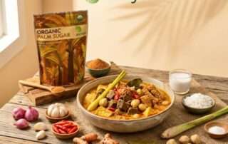 Sebuah foto makanan estetik yang menangkap mangkuk besar hidangan resep Bumbu Rujak campur Banyumas yang berisi daging sapi, tahu goreng, dan tempe goreng dalam kuah santan kental yang kaya warna, dihiasi serai, daun salam, dan cabai merah iris. Hidangan ini dikelilingi oleh bahan-bahan baku mentah: bawang merah, bawang putih, cabai rawit merah, kunyit, daun salam, serai, garam, mangkuk kelapa parut, dan gelas santan cair. Di sisi kiri berdiri kemasan Arenga Organic Palm Sugar 225g dengan semangkuk kecil gula bubuk dan sendok kayu. Meja kayu pedesaan dengan latar belakang krem hangat dan bayangan daun palem.