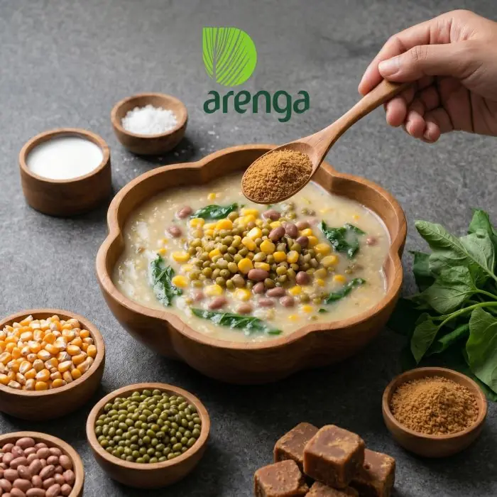 Penyajian hidangan Jagung Bose Timor yang kental dan bergizi dalam mangkuk kayu berbentuk bunga, dengan tangan menuangkan gula aren organik Arenga dari sendok, disajikan di atas permukaan batu bersama bahan-bahan mentah sebagai alternatif sarapan sehat.