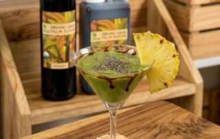 Segelas minuman hijau segar dari Resep Smoothies untuk Kecantikan Kulit dengan taburan chia seed, hiasan potongan nanas, dan lelehan gula aren cair Arenga di dalam gelas. Disajikan di atas tatakan bambu pada meja kayu dengan latar belakang kemasan Arenga Organic Liquid Palm Sugar.