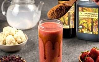 Segelas resep smoothie strawberry rosela yang menyegarkan dengan lelehan gula aren cair Arenga di dinding gelas, disajikan berdampingan dengan botol Arenga Organic Liquid Palm Sugar dan stroberi segar sebagai minuman tropis penjaga awet muda