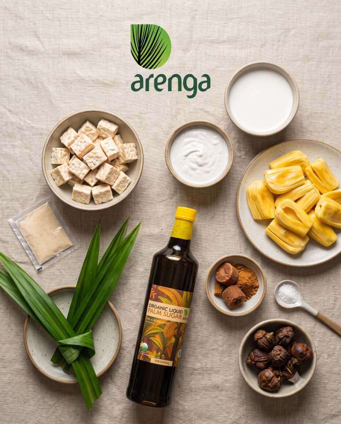 Foto flat-lay bahan-bahan resep puding tempe diatur rapi di atas permukaan kayu bertekstur. Mangkuk-mangkuk berisi tempe kukus halus, potongan dadu nangka kuning matang, santan ganda (kental dan encer), dan paket agar-agar bubuk putih. Botol Arenga gula aren cair, garam, dan ikatan daun pandan segar melengkapi komposisi visual panduan resep.