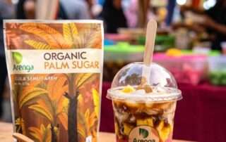 Segelas koktail mutiara kentang gula aren untuk jualan dalam cup plastik transparan, menampilkan lapisan ketan hitam, sagu mutiara pink, dan potongan nangka bersiram santan. Di sebelahnya terdapat kemasan Arenga Organic Palm Sugar dan mangkuk kaca berisi gula aren bubuk, dengan latar belakang lapak takjil yang diburamkan.