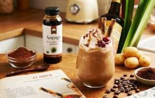 Foto studio yang estetis di atas meja kayu pedesaan. Di tengah adalah gelas tinggi berisi frappe moka kentang ungu berlapis dengan krim kocok kaya, gerimis sirup cokelat, taburan kakao, dan keripik kentang ungu renyah sebagai hiasan. Di latar belakang, terdapat kentang ungu utuh dan belah, serta mangkuk kecil berisi gula aren blok dan sebotol kecil Arenga Gula Aren Cair di sebelah kiri.
