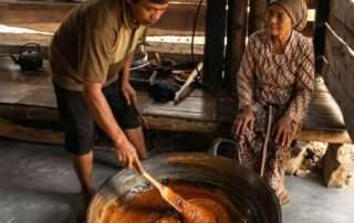 Sepasang suami istri perajin sedang mengaduk nira kental di kuali besar dalam dapur tradisional yang bersih. Visual ini mewakili cerita sepasang suami istri perajin gula aren, sebuah rahasia storytelling manis untuk bisnis kafe dan bakery.